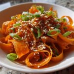 Close-up of a vibrant raw carrot salad featuring shaved carrot ribbons, topped with sesame seeds and sliced green onions.