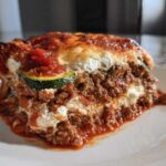A thick, layered slice of zucchini lasagna featuring meat sauce, ricotta cheese, and a visible zucchini noodle layer.