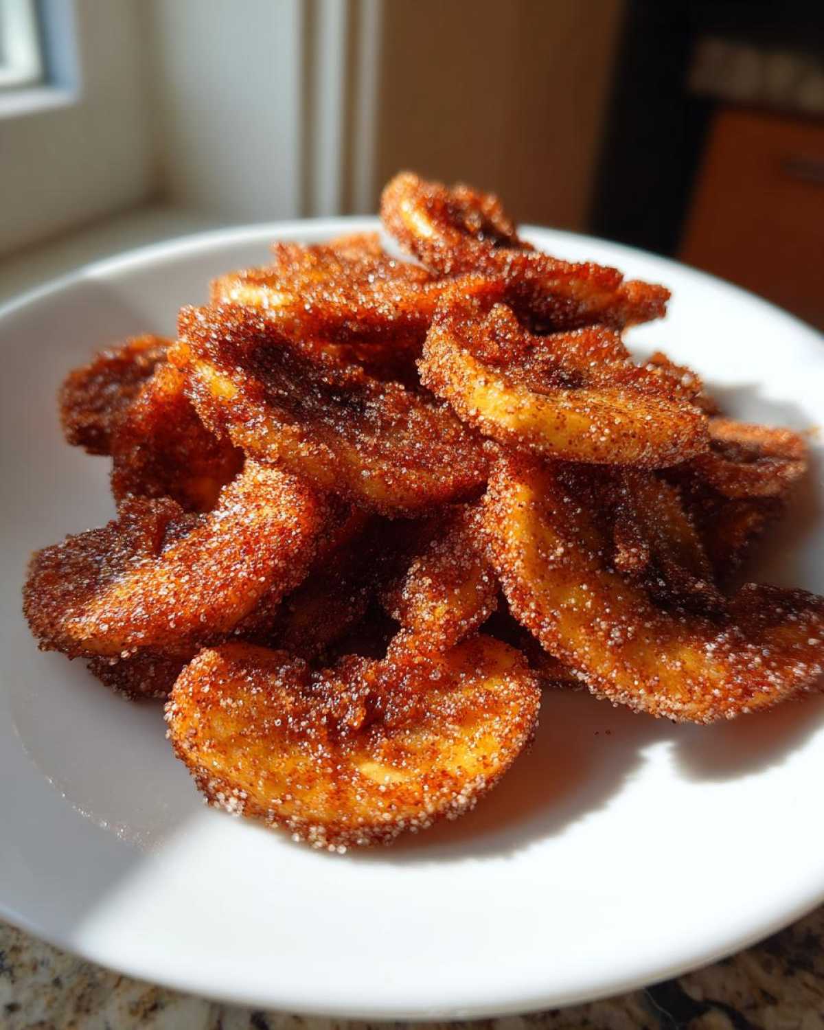 A pile of Air Fryer Crispy Cinnamon Sugar Banana Chips coated heavily in cinnamon sugar on a white plate.