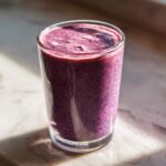 A tall glass filled with a thick, vibrant purple Creamy Blueberry Banana Smoothie, sitting on a marble surface.