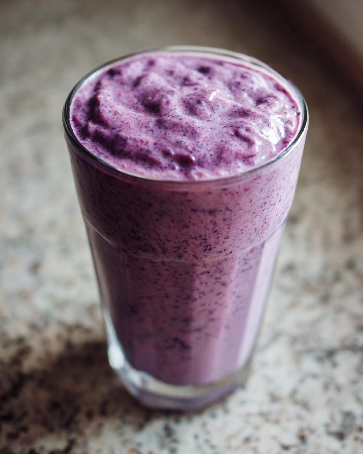 Close-up of a thick, purple Creamy Blueberry Banana Smoothie filling a tall glass on a speckled countertop.