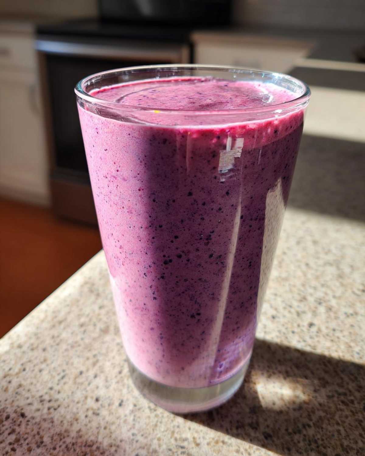 A tall glass filled with a thick, vibrant purple Creamy Blueberry Banana Smoothie sitting on a speckled countertop.