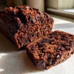 A dark, rich loaf of Gluten-Free Chocolate Zucchini Bread with a slice cut off, showing moist texture and chocolate chips.