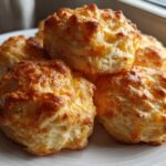 A close-up of several golden-brown Gluten-Free Garlic Cheddar Biscuits piled high on a white plate.