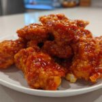 A pile of crispy chicken pieces coated in a thick, glossy, orange-red Honey Sriracha Chicken glaze on a white plate.