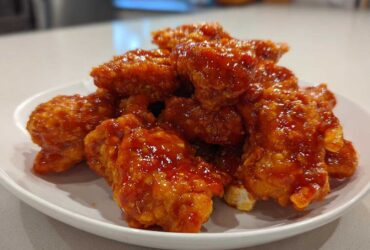 A pile of crispy chicken pieces coated in a thick, glossy, orange-red Honey Sriracha Chicken glaze on a white plate.