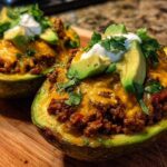 Two halves of Keto Taco Stuffed Avocados filled with seasoned ground meat, melted cheese, and topped with sour cream, avocado slices, and cilantro.