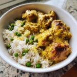 Close-up of a white bowl filled with One-Pot Coconut Curry Chicken and Rice, featuring creamy yellow curry sauce and bright green peas.