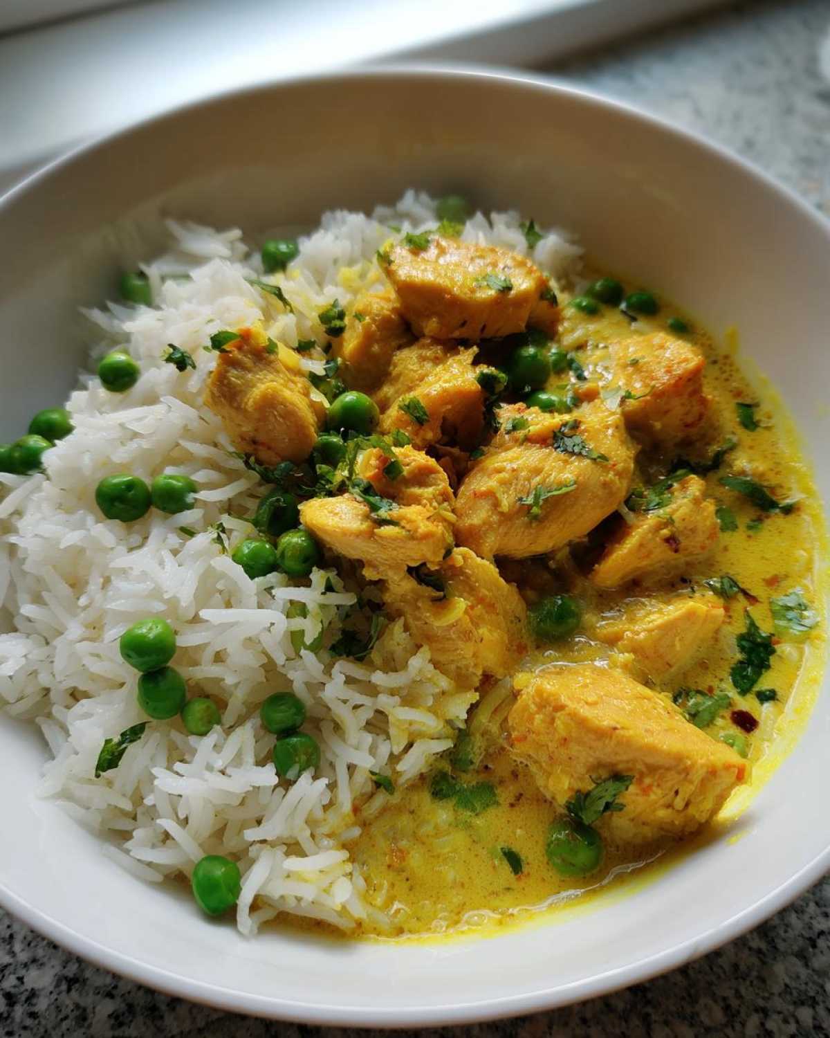 Close-up of a white bowl filled with One-Pot Coconut Curry Chicken and Rice, featuring yellow curry sauce, chicken pieces, white rice, and green peas.
