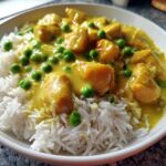 Close-up of a white bowl filled with fluffy white rice topped with creamy yellow coconut curry chicken and bright green peas.