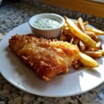 A plate featuring golden, crispy Copycat Pub-Style Fish and Chips served with a side of homemade tartar sauce.