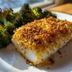 Close-up of a flaky white fish fillet with a golden Sheet Pan Parmesan Crusted Cod topping next to roasted broccoli.