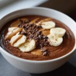 Close-up of a thick Vegan Chocolate Peanut Butter Banana Smoothie Bowl topped with banana slices, peanut butter swirls, and cacao nibs.