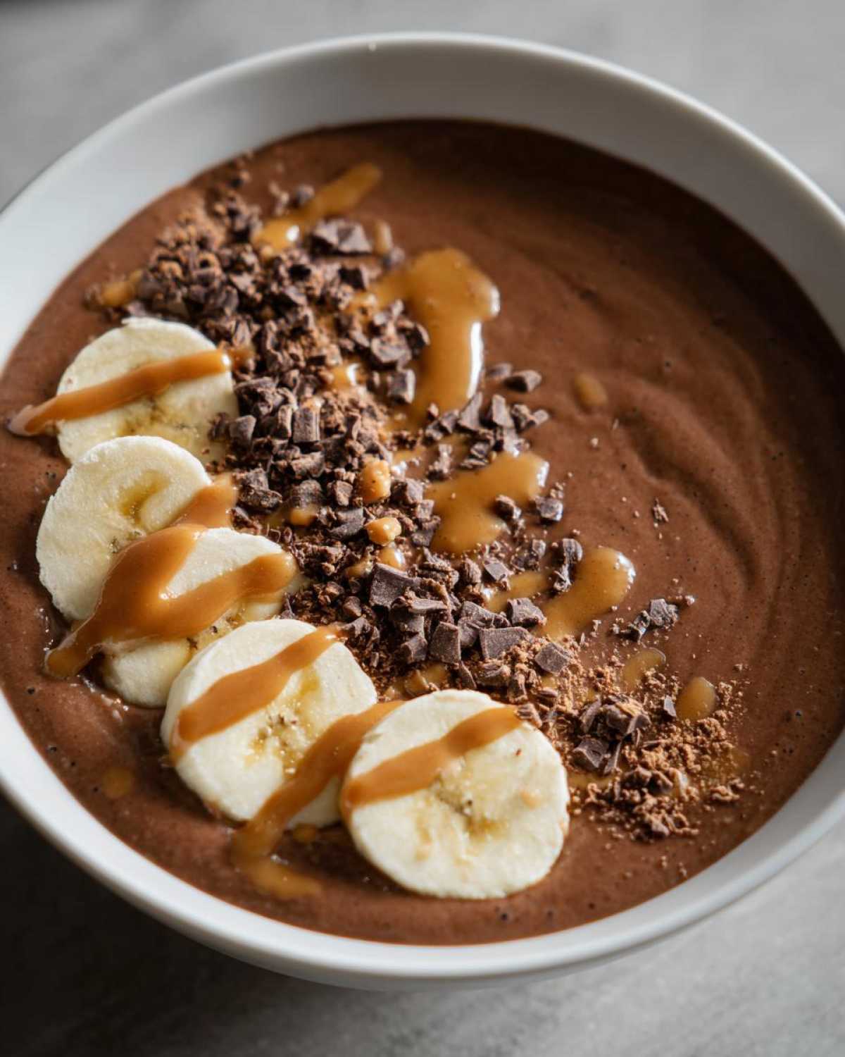 Close-up of a thick Vegan Chocolate Peanut Butter Banana Smoothie Bowl topped with banana slices, chocolate shavings, and peanut butter drizzle.