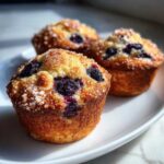 Three golden-brown Vegan Lemon Blueberry Muffins topped with coarse sugar crystals and bursting blueberries.