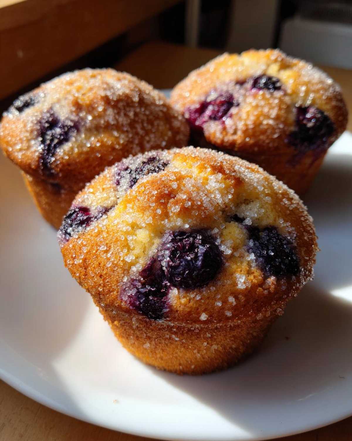 Three golden-brown Vegan Lemon Blueberry Muffins topped with coarse sugar crystals, sitting on a white plate.