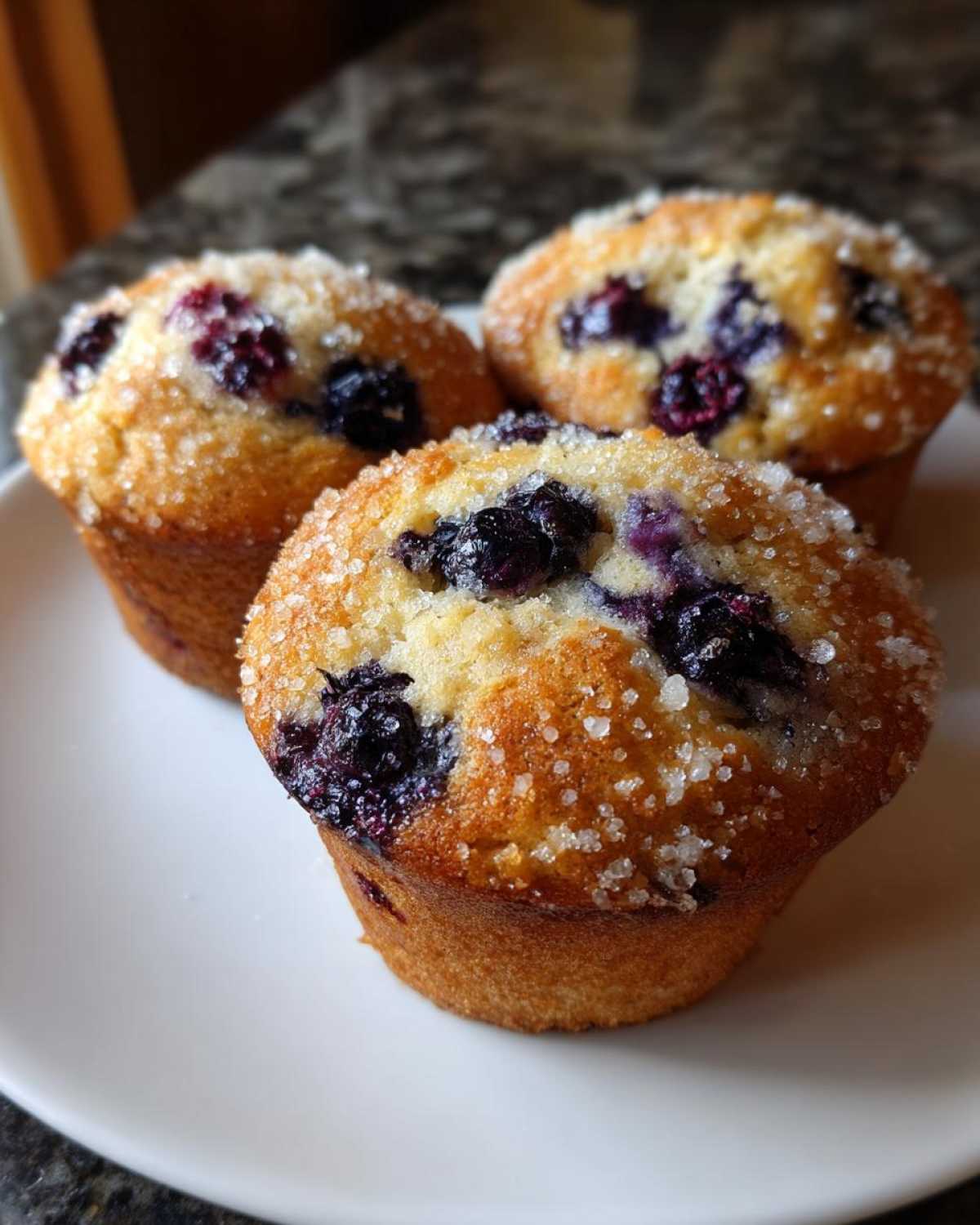 Three golden-brown Vegan Lemon Blueberry Muffins topped with coarse sugar crystals, sitting on a white plate.