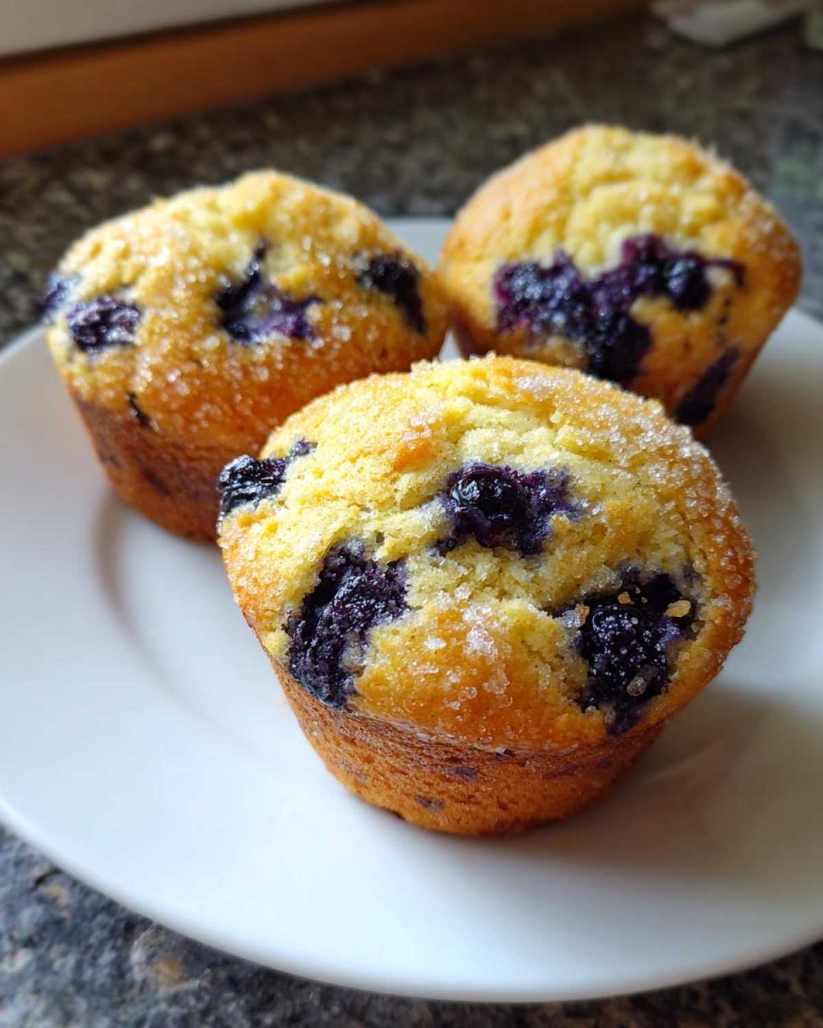 Three golden-brown Vegan Lemon Blueberry Muffins topped with coarse sugar crystals, served on a white plate.