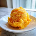 A bright orange scoop of textured Vegan Mango Pineapple Sorbet served on a small white dish.