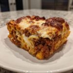 A generous square slice of Cheesy Baked Ziti with Ground Beef and Sauce, showing layers of pasta, meat sauce, and melted browned cheese topping.
