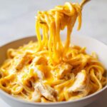 A fork lifting long strands of pasta coated in bright orange, creamy sauce from a bowl of Cheesy Chicken Pasta with Creamy Sauce.