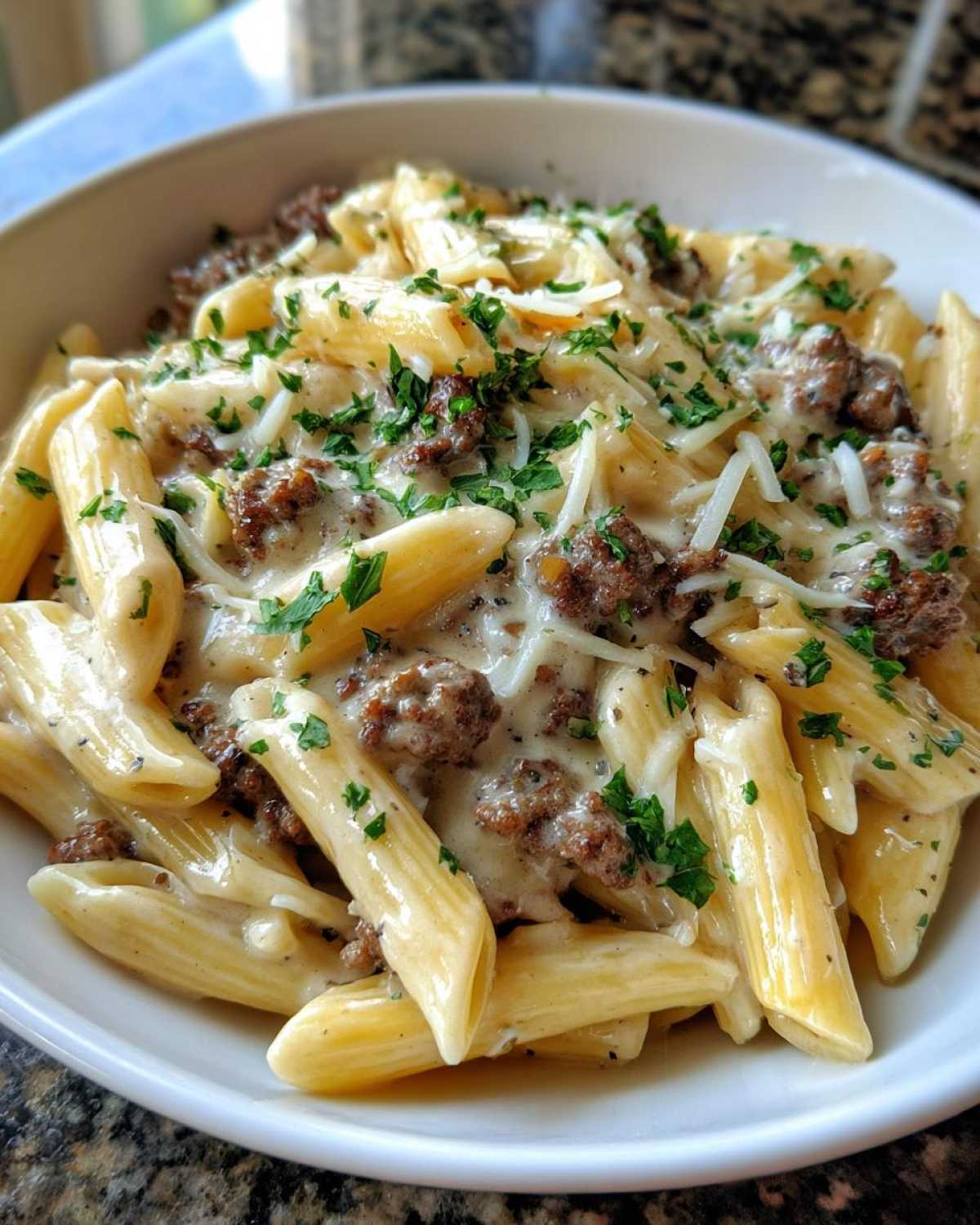 A close-up of a white bowl filled with Cheesy Ground Beef Pasta with Garlic Cream Sauce, topped with shredded cheese and parsley.