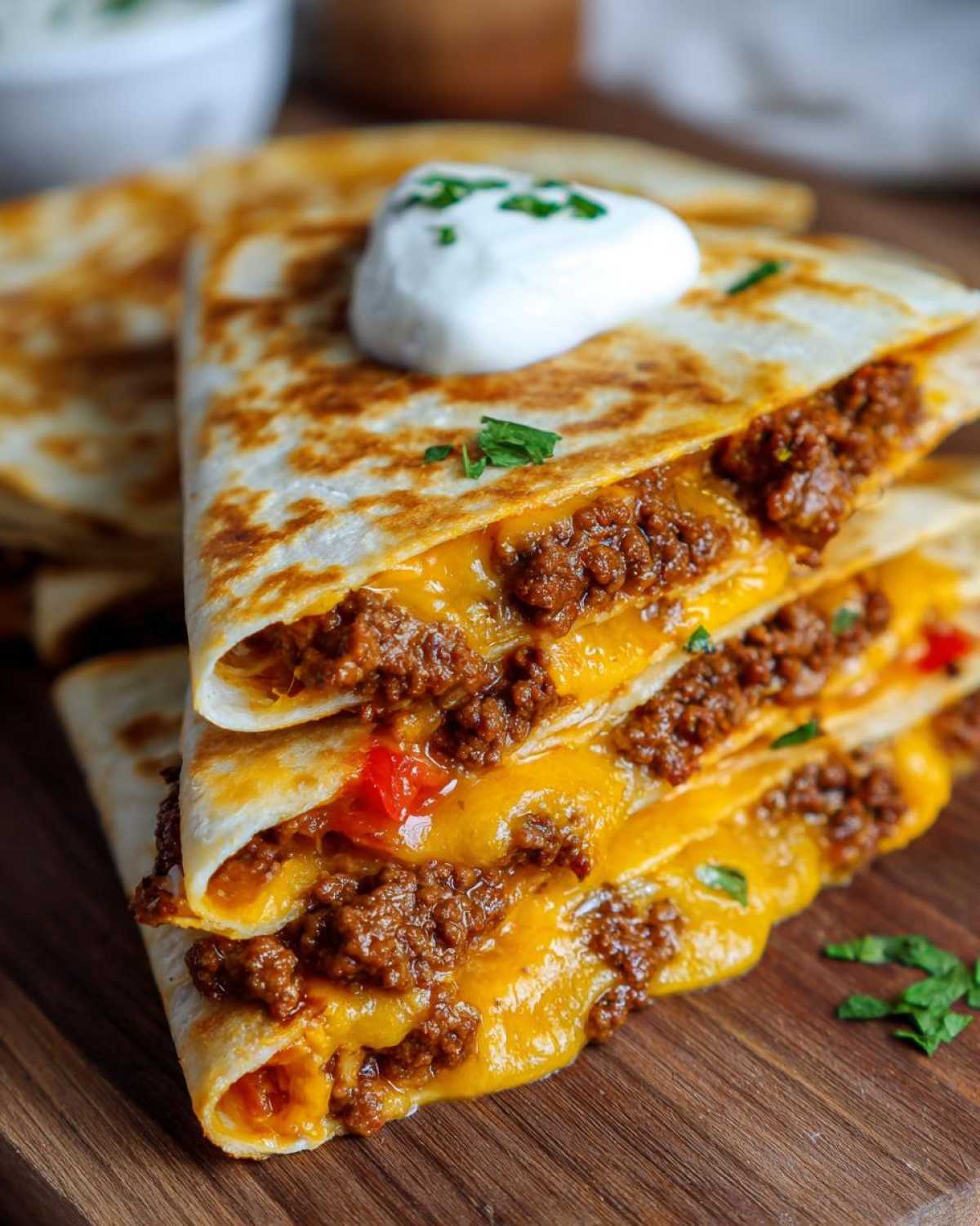 Stack of three cheesy ground beef quesadilla wedges topped with sour cream and parsley.