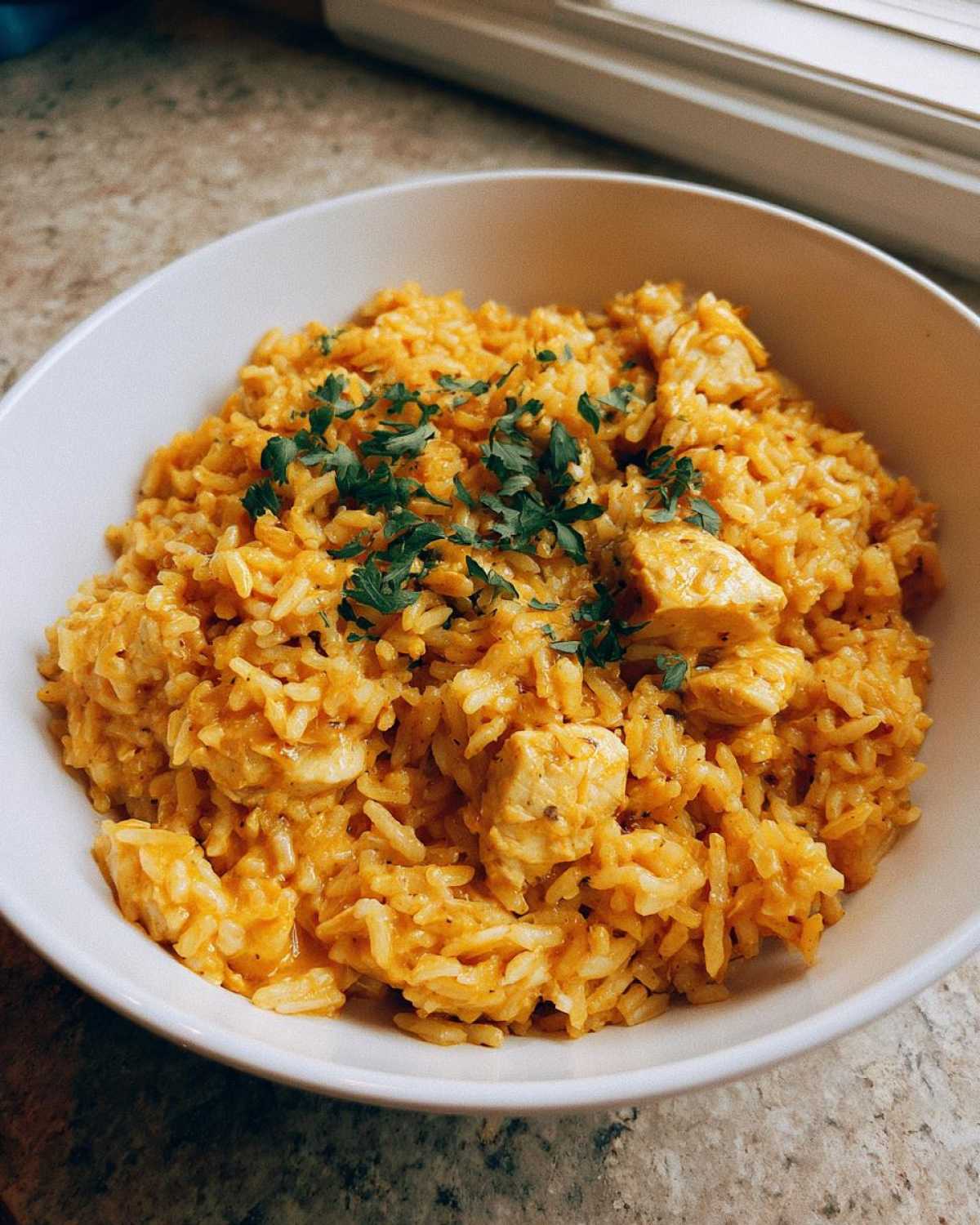 A white bowl filled with a rich, orange-hued Creamy Cajun Chicken Rice One Pot Meal, topped with fresh parsley.