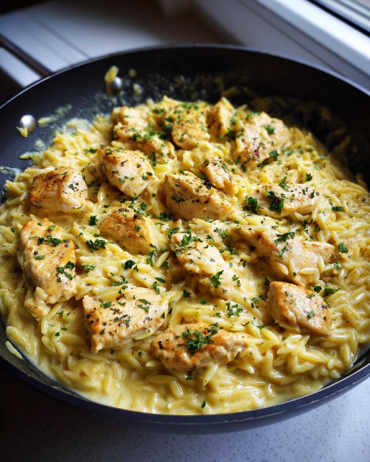 Close-up of Creamy Garlic Parmesan Orzo with Chicken pieces garnished with parsley in a black skillet.
