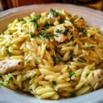 A close-up of a bowl of Creamy Lemon Chicken Orzo, featuring rice-shaped pasta, chunks of chicken, and fresh green herbs.