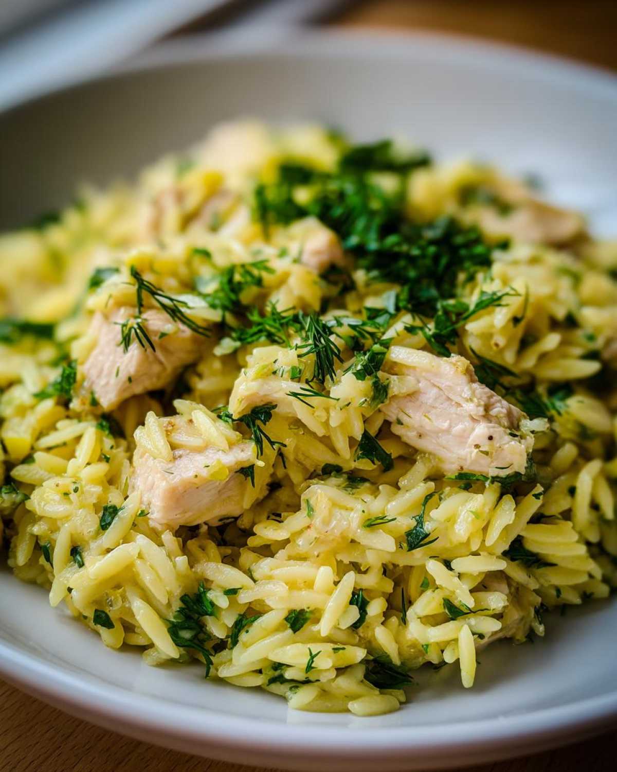 A close-up, appetizing shot of creamy lemon chicken orzo pasta mixed with chunks of chicken and topped generously with fresh green herbs.