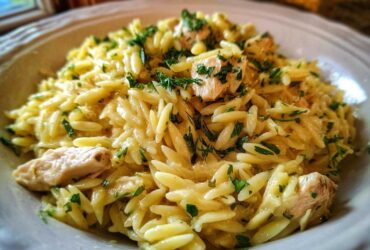 A close-up of a bowl of Creamy Lemon Chicken Orzo, featuring rice-shaped pasta, chunks of chicken, and fresh green herbs.