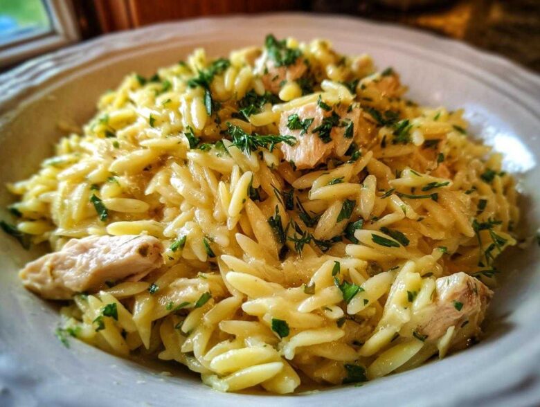 A close-up of a bowl of Creamy Lemon Chicken Orzo, featuring rice-shaped pasta, chunks of chicken, and fresh green herbs.