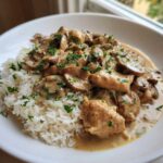 A close-up of a white bowl filled with white rice topped with creamy mushroom chicken and garnished with fresh parsley.