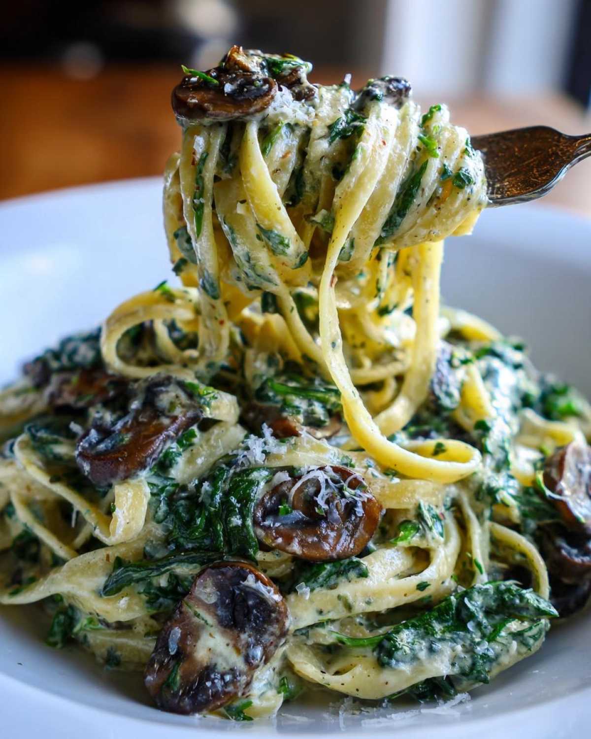 A fork lifts fettuccine coated in rich sauce, spinach, and mushrooms from a bowl of Creamy Spinach and Mushroom Pasta Dinner.