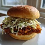 A close-up of a Crispy Chicken Sandwich with Creamy Slaw piled high on a toasted bun, sitting on a white plate.