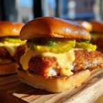 A close-up of a Crispy Chicken Slider featuring a golden fried patty, melted cheese sauce, and pickles on a brioche bun.