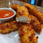 Golden brown, crispy chicken strips served with a small bowl of bright orange spicy dip.