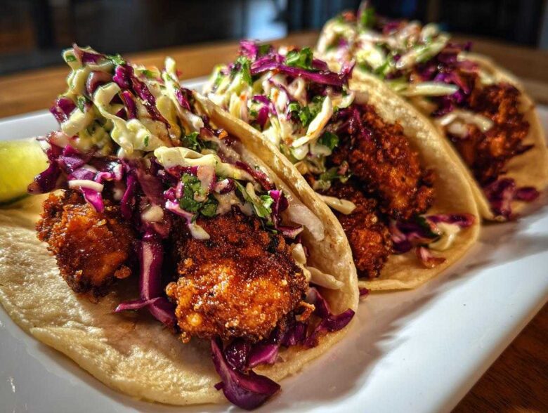 Three Crispy Chicken Tacos filled with crunchy fried chicken, vibrant purple and green slaw, served on a white plate.