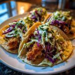 Four Crispy Chicken Tacos topped generously with creamy slaw and cilantro sauce on a white plate.