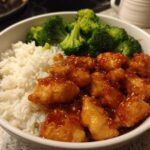 A close-up of a white bowl filled with Honey Garlic Chicken Bowl with Rice, topped with steamed broccoli florets.