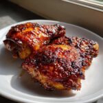 Three perfectly glazed Honey Garlic Chicken Thighs Oven Baked pieces resting on a white plate, showing caramelized, sticky skin.