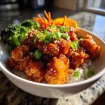 A close-up of a Honey Sriracha Chicken Bowl featuring glazed chicken pieces over rice, topped with sesame seeds and green onions.