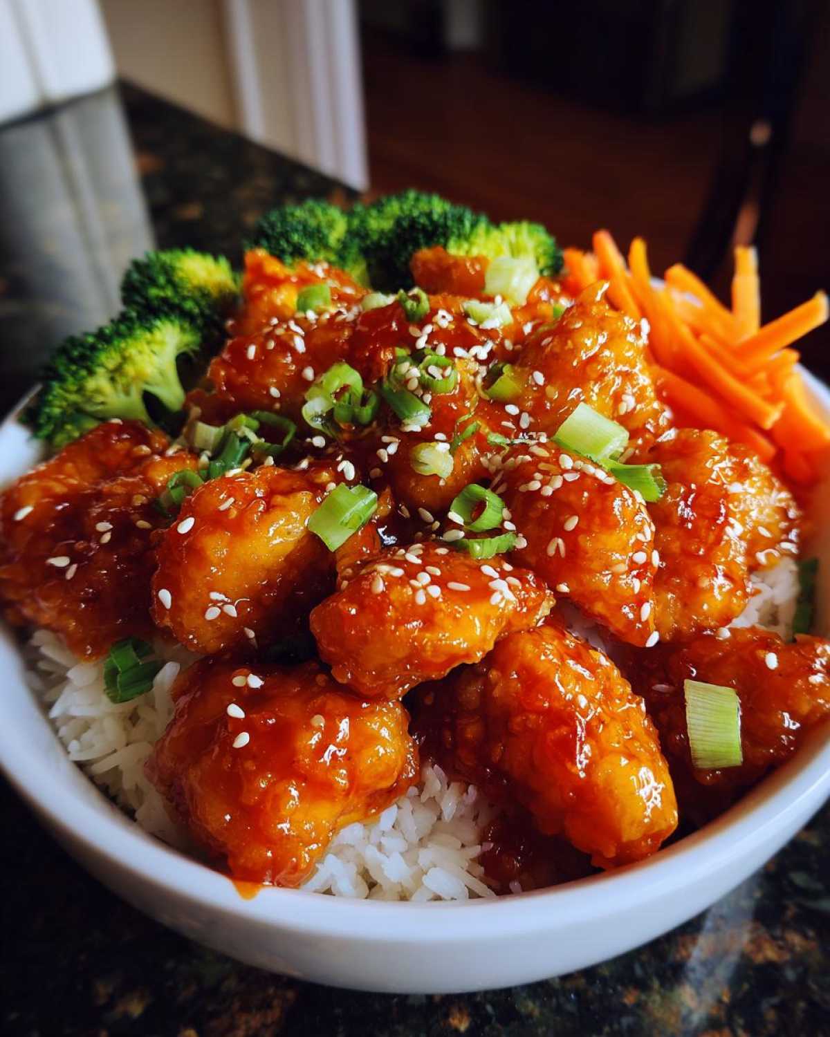 A close-up of a Honey Sriracha Chicken Bowl featuring glazed chicken pieces over rice, topped with sesame seeds and green onions, served with broccoli and carrots.