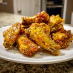A pile of golden, crispy Lemon Pepper Chicken Wings Oven Style seasoned heavily with black pepper, served on a white plate.