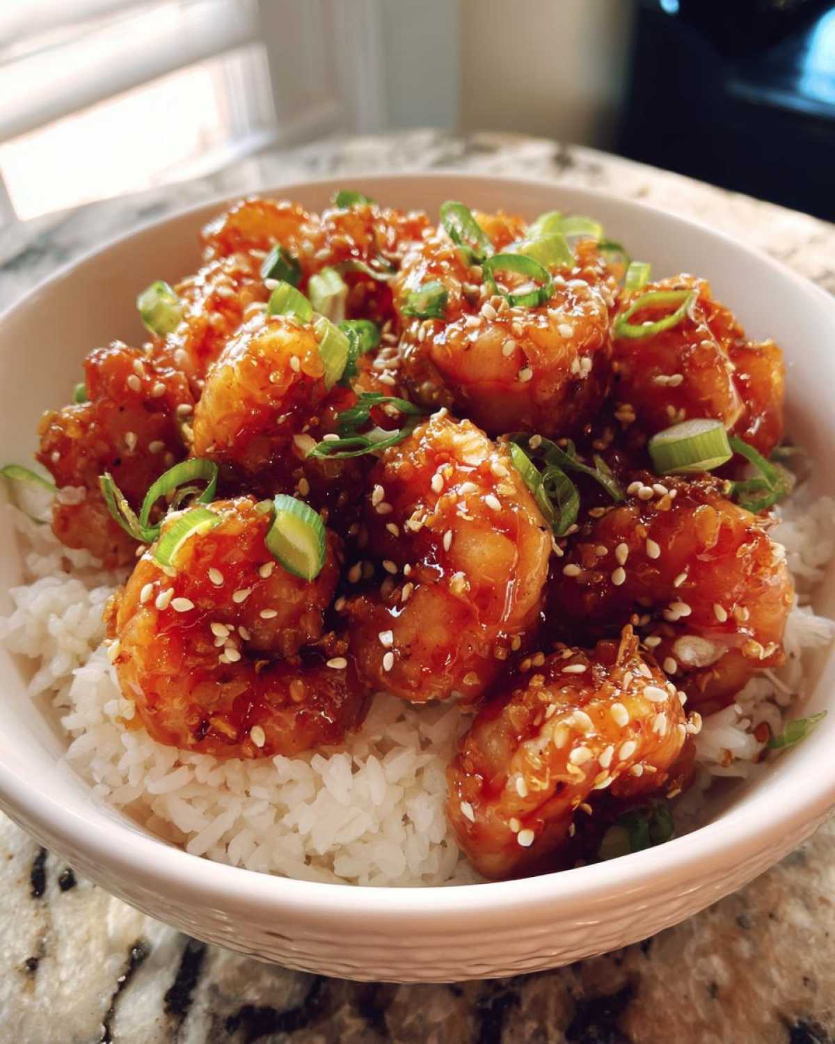 Close-up of a white bowl filled with white rice topped with glossy Spicy Honey Garlic Shrimp and garnished with sesame seeds and green onions.