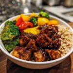 A white bowl filled with Sticky BBQ Chicken Bowl with Veggies, featuring saucy chicken pieces over brown rice and colorful bell peppers and broccoli.