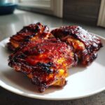 Three glazed Sticky BBQ Chicken Thighs with crispy, caramelized edges served on a white plate.