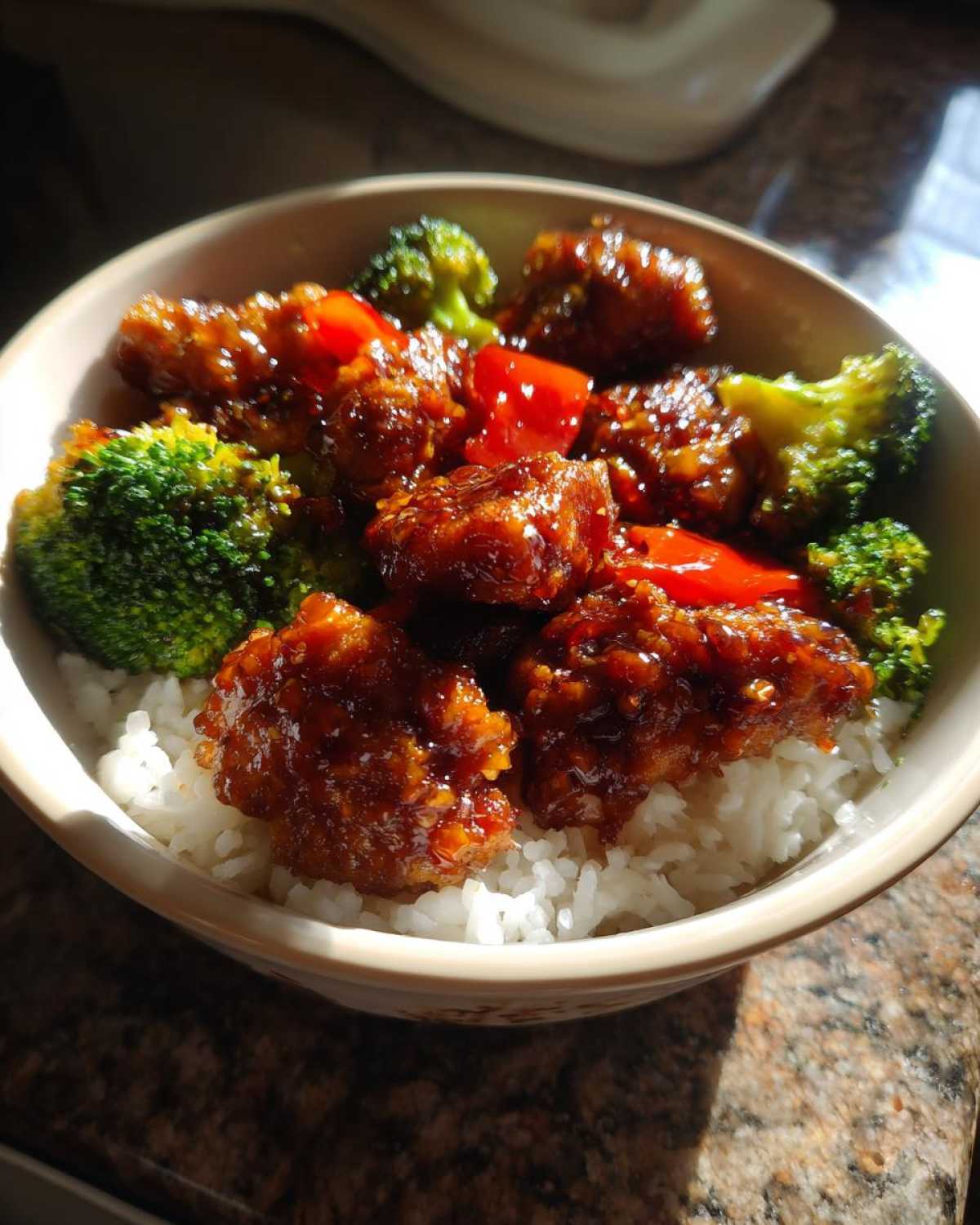 A close-up of a Sticky Orange Chicken Bowl with rice, vibrant broccoli florets, and red peppers coated in a glossy sauce.