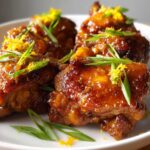 Close-up of four glazed Sticky Orange Chicken Thighs Better Than Takeout garnished with green onion and orange zest.